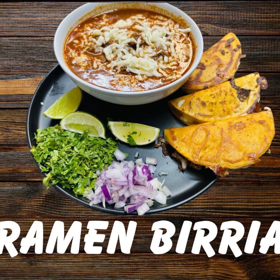 Birria ramen with cheese, birria tacos, cilantro, onions, and lime wedges on a dark plate.