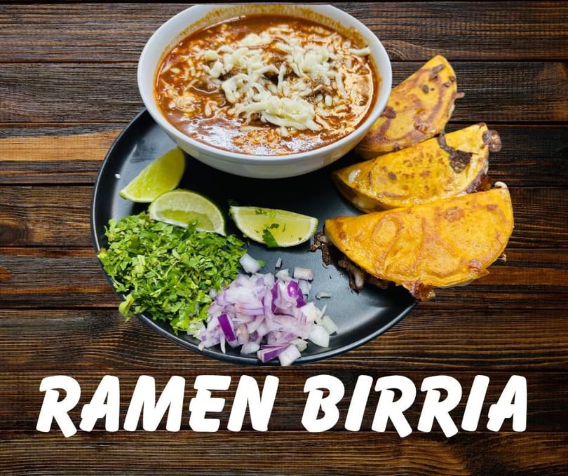 Birria ramen with cheese, birria tacos, cilantro, onions, and lime wedges on a dark plate.